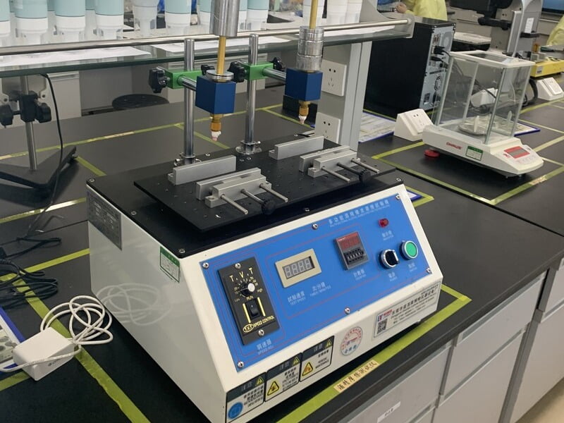 Cosmetic packaging testing equipment with a control panel and laboratory balance on a workbench.